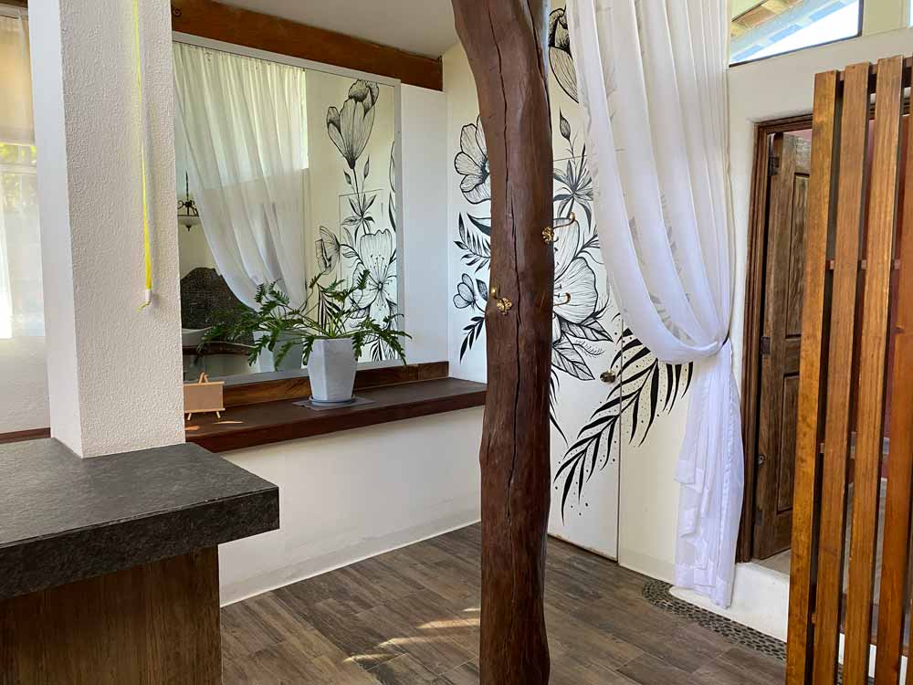 Una habitación serena con detalles de madera cuenta con un gran espejo, una planta en maceta y un mural de pared con diseños florales. Cortinas blancas transparentes cuelgan cerca de una puerta de madera y la luz natural entra a raudales, resaltando el piso de madera y una viga de madera vertical.