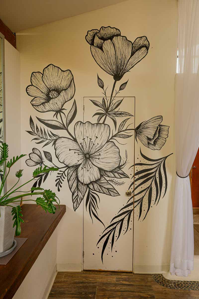 Arte mural sobre una puerta con grandes ilustraciones florales en blanco y negro con hojas y flores, sobre una pared de color crema. A la derecha cuelga una cortina blanca transparente y a la izquierda se ve parte de una encimera de madera con plantas verdes.