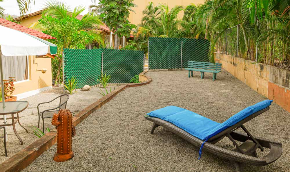 Patio cubierto de grava con un sillón con cojines azules, un banco de madera, una cerca verde y plantas tropicales. Cerca hay una mesa redonda con sillas y un paragüero. A la izquierda hay una boca de incendios de color óxido.