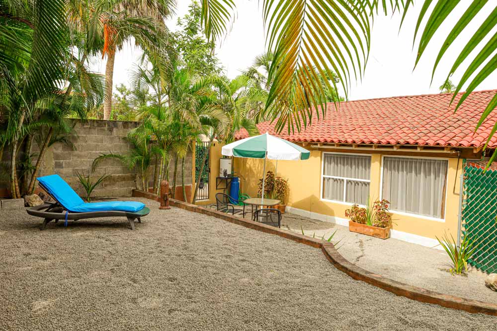 Un tranquilo patio trasero con una pequeña casa con techo de tejas rojas. Hay un sillón azul sobre grava, rodeado de vegetación exuberante. Cerca de la casa hay una mesa de patio con sillas y una sombrilla, lo que crea un acogedor espacio al aire libre.