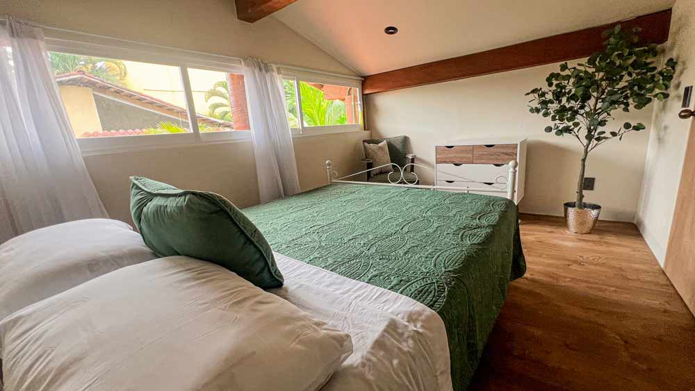 Acogedor dormitorio en bungalow cerca de Playa La Ropa en Zihuatanejo, con un cubrecama verde, almohadas verdes y blancas y una cómoda blanca y de madera. Una planta en maceta se encuentra en la esquina mientras la luz natural se cuela a través de grandes ventanales con cortinas transparentes, que resaltan los pisos y las vigas de madera.