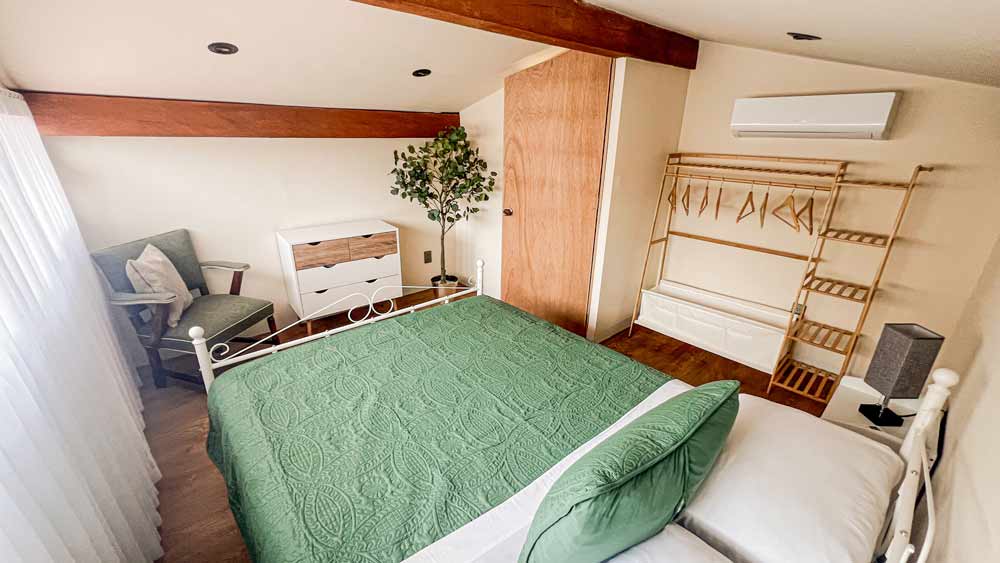 Un dormitorio acogedor en un bungalow cerca de Playa La Ropa, con una cama acolchada verde, vigas de madera y paredes claras. Una pequeña cómoda y una planta en maceta se encuentran junto a una ventana con cortinas transparentes. Un estante de madera con perchas y un cojín de silla brindan encanto, mientras que un aire acondicionado garantiza comodidad en el calor de Zihuatanejo.