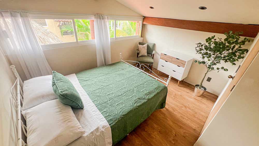 Acogedor dormitorio en un encantador bungalow cerca de Playa La Ropa, con una cama cuidadosamente tendida con un cubrecama y almohadas verdes. Una pequeña cómoda, una planta en maceta y una silla acolchada adornan el espacio. La luz del sol se filtra a través de las cortinas transparentes de las largas ventanas horizontales, lo que genera un brillo cálido en todo el lugar.