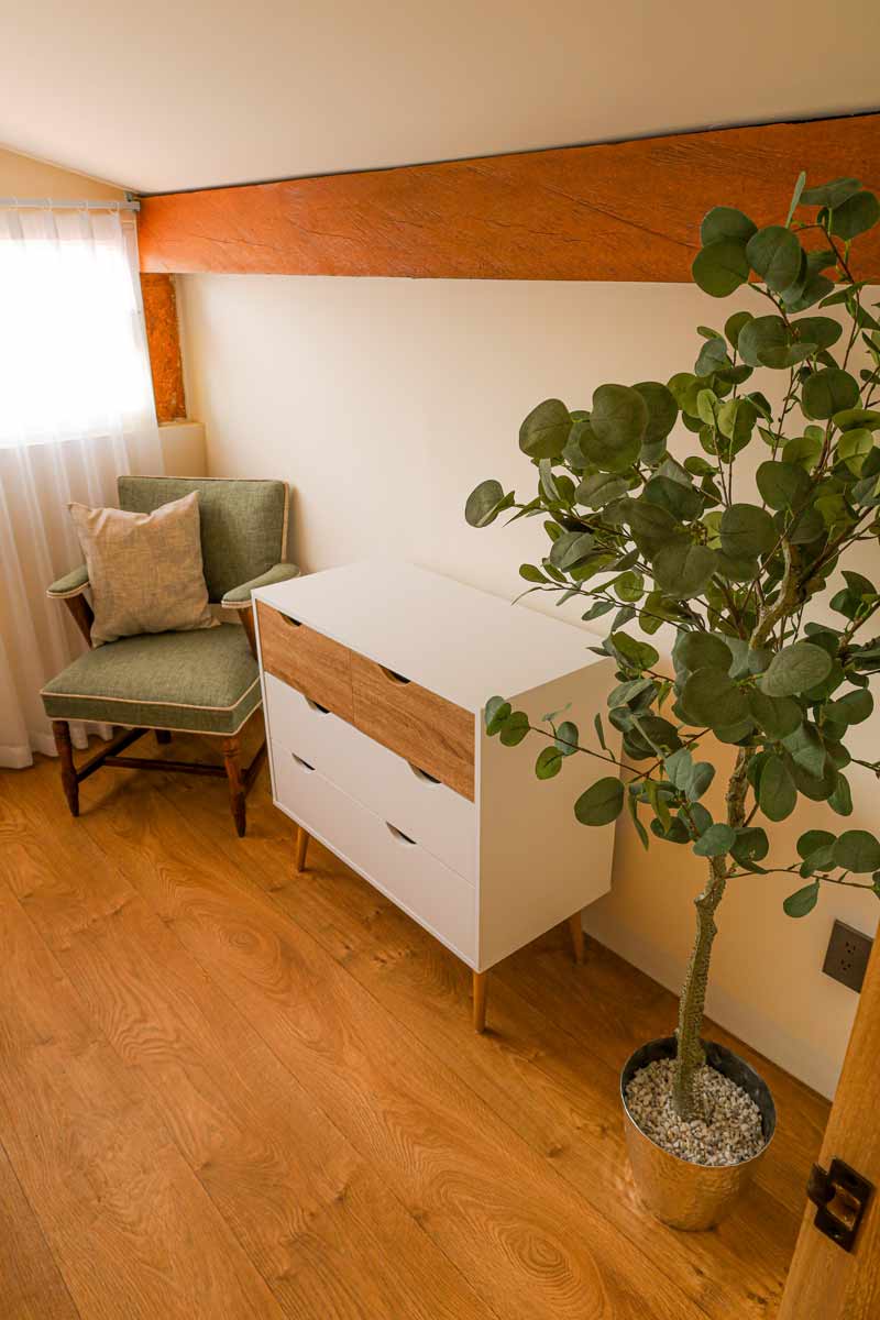 En un rincón acogedor que recuerda a un bungalow cerca de Playa La Ropa, la habitación cuenta con un piso de madera, una silla acolchada verde con una almohada beige y una cómoda blanca con cajones de madera. Una planta en maceta crece mientras la luz suave se filtra a través de una cortina transparente a la izquierda.